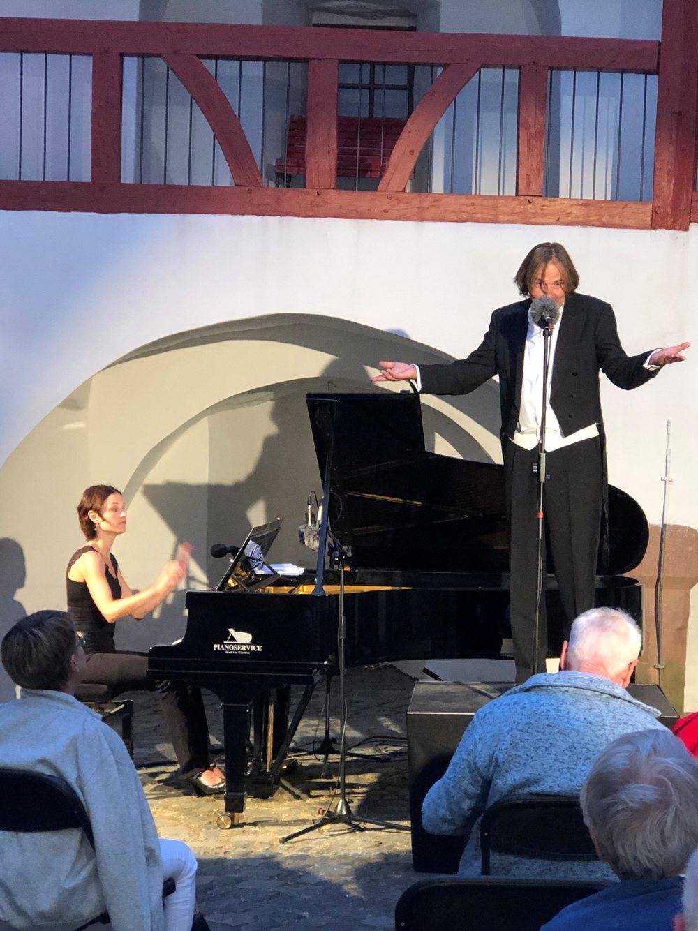 Ein Sänger im Frack breitet vor weißen Arkaden die Arme aus, am Flügel eine Pianistin, davor Zuhörer