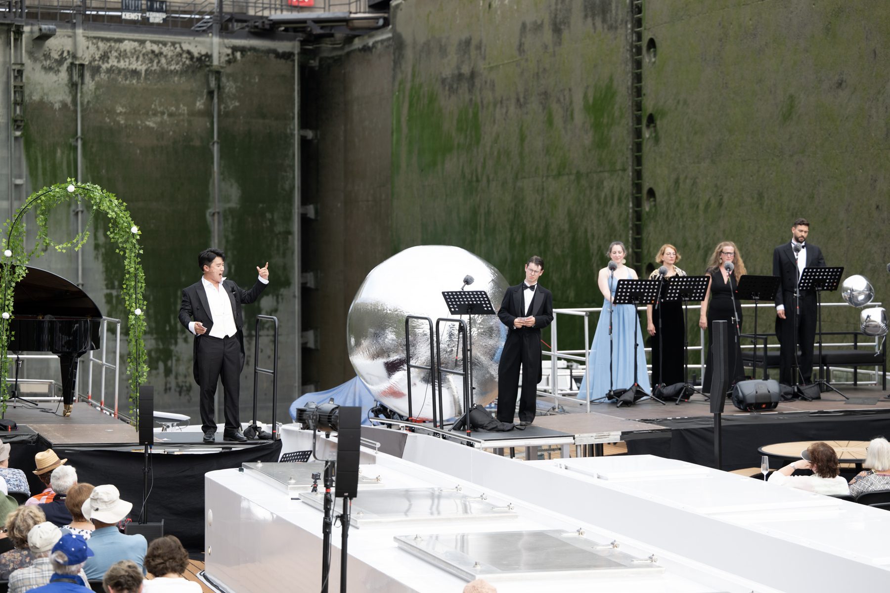 Das Ensemble auf dem Deck: ein Sänger im Frack, mehrere Sängerinnen und Sänger mit Notenpulten, daneben eine große silberne Kugel