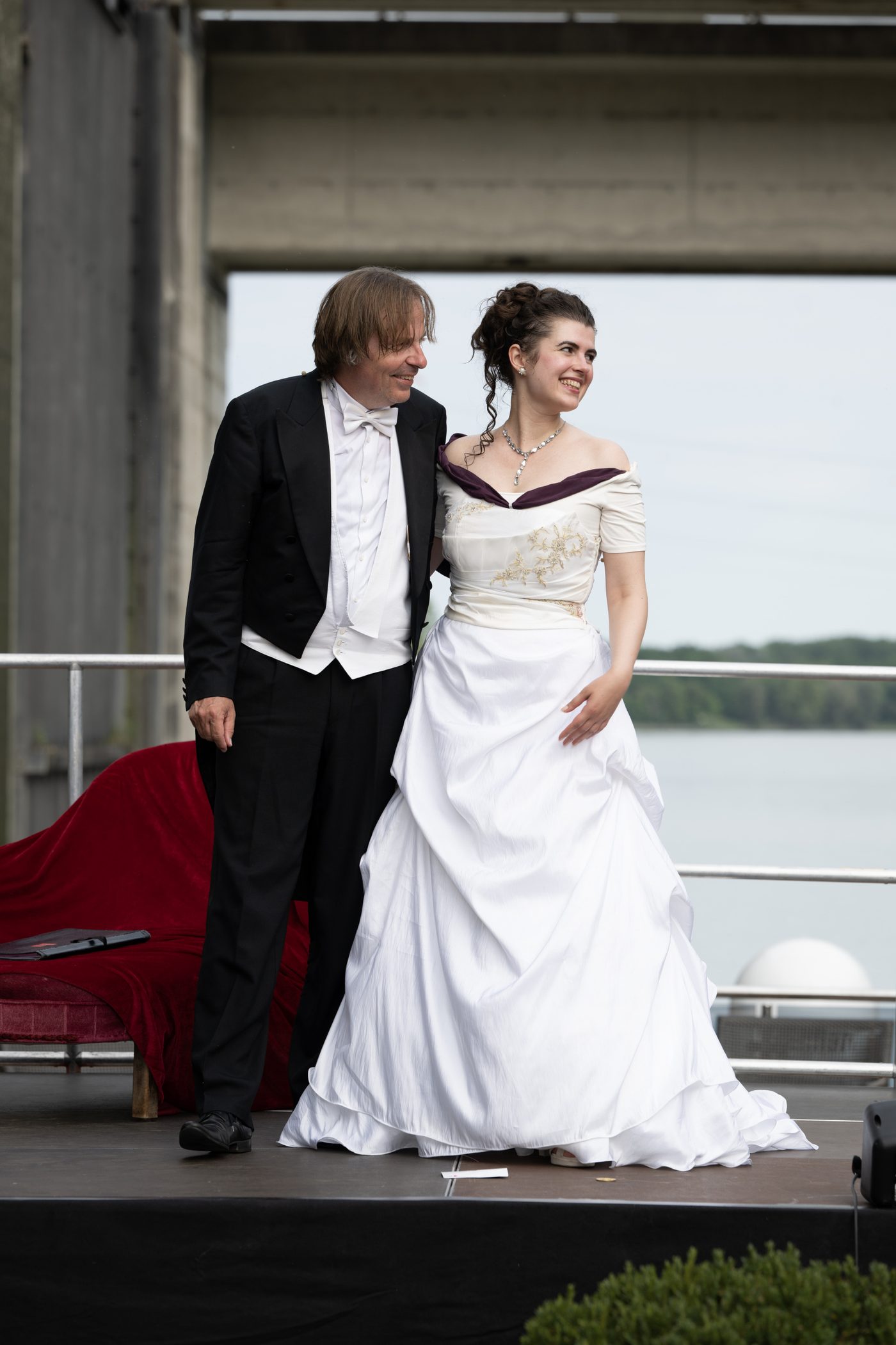 Ein Sänger im Frack und eine Sängerin im weißen Kleid stehen lächelnd nebeneinander, hinter ihnen der Fluss