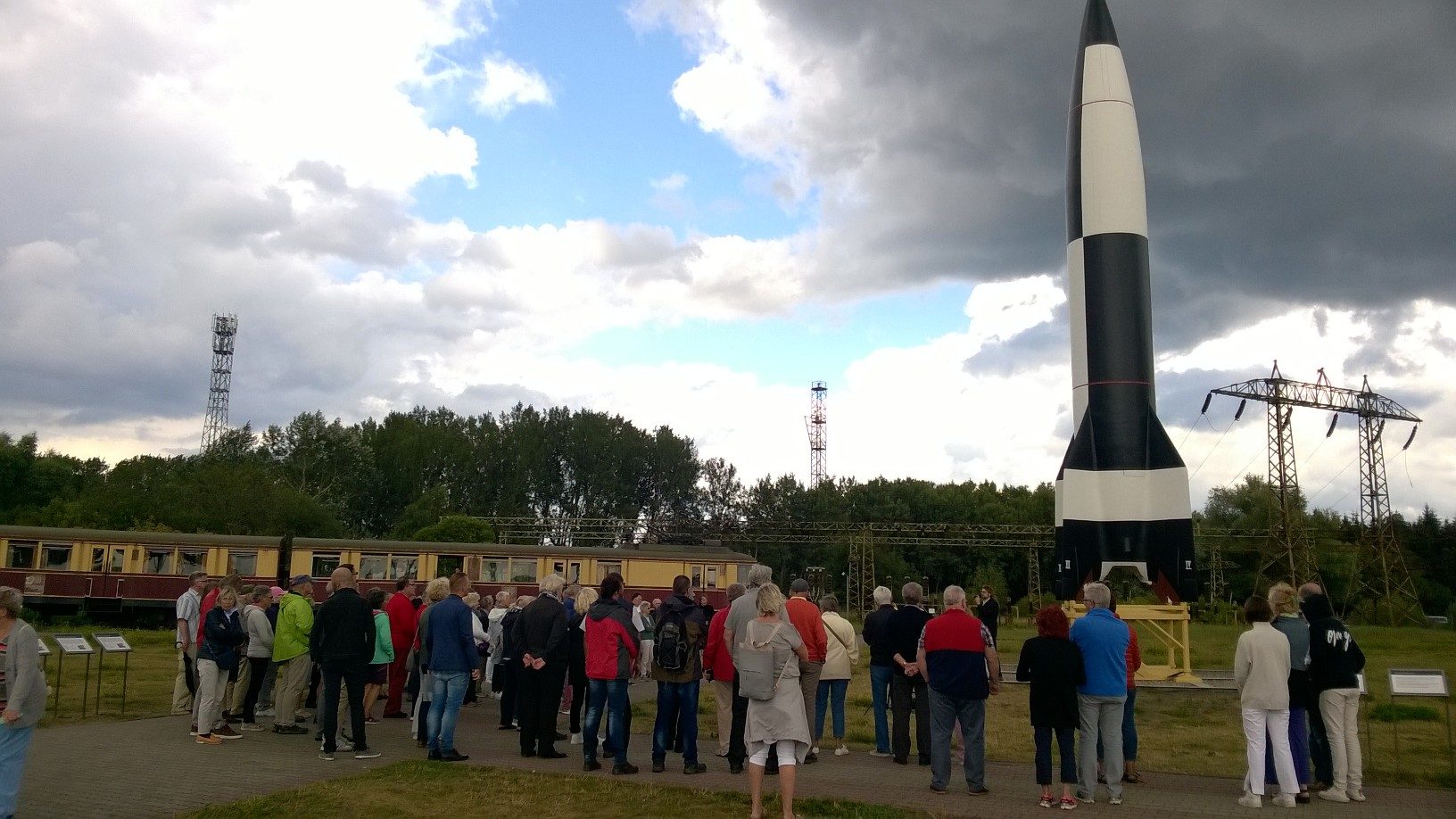 Besucher stehen vor einer schwarz-weißen Rakete auf dem Museumsgelände, dahinter Bäume und Strommasten