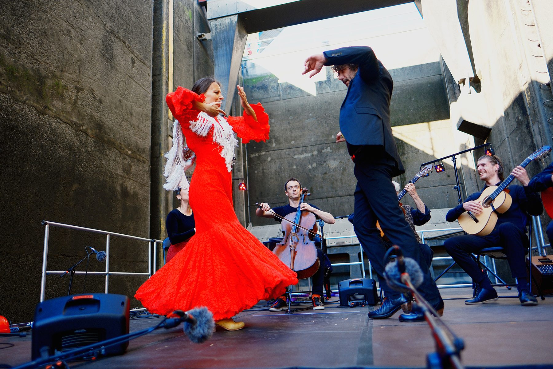 Eine Flamenco-Tänzerin im roten Kleid und ein Tänzer in Schwarz tanzen vor Gitarristen und einer Cellistin vor der Schleusenwand