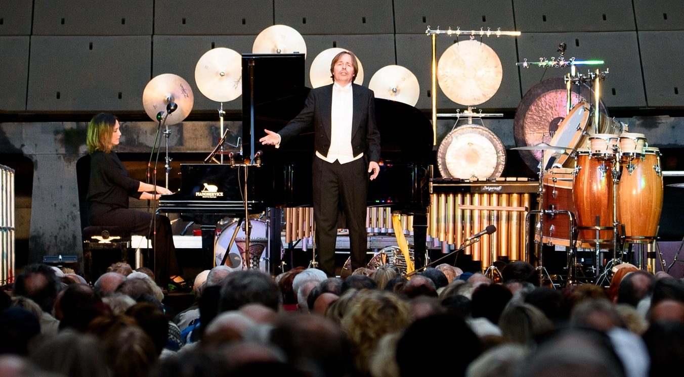 Ein Sänger im Frack breitet auf der Bühne die Arme aus, links eine Pianistin am Flügel, dahinter Becken und Gongs