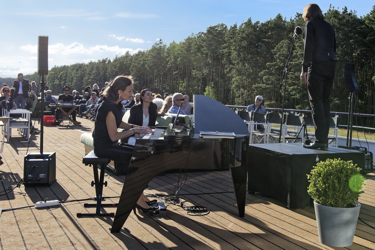 Eine Pianistin am Flügel und ein Sänger auf dem Sonnendeck, dahinter Zuhörer und Kiefernwald am Kanal
