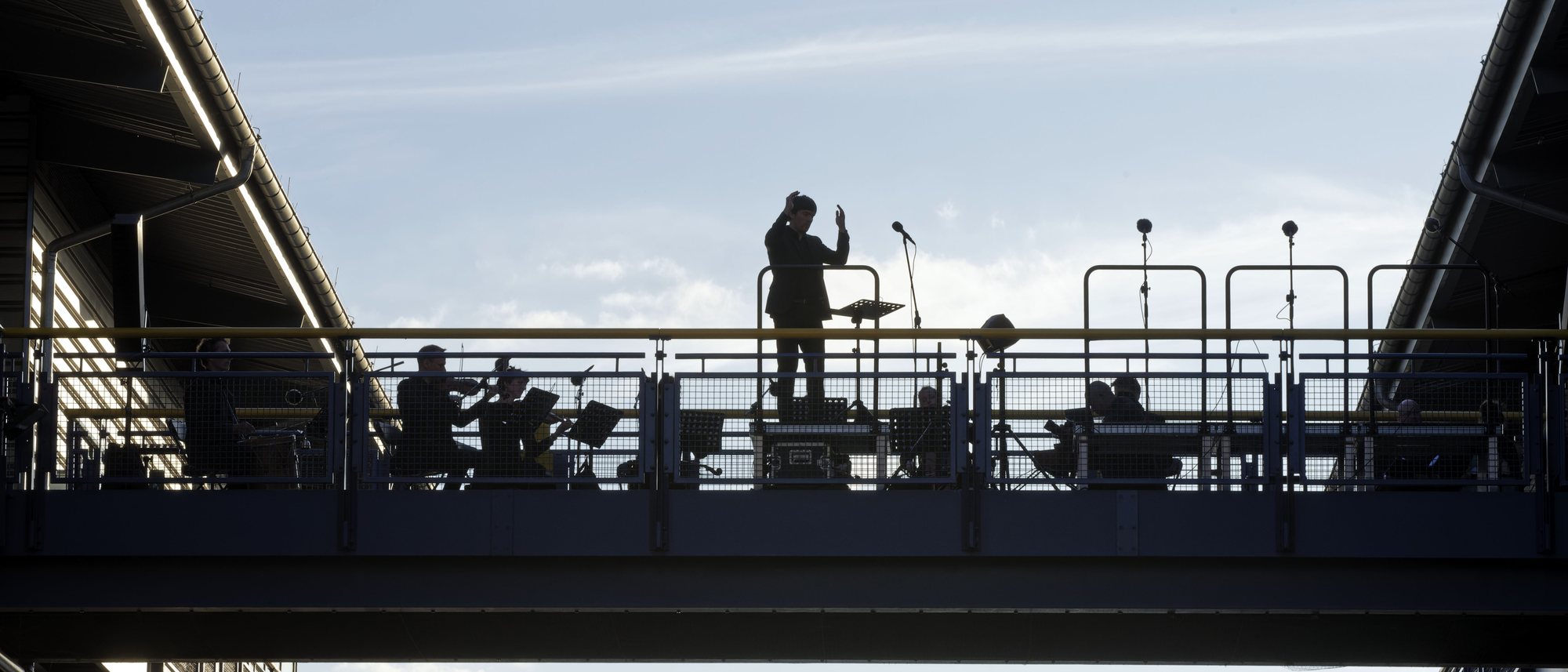 Ein Dirigent als Scherenschnitt auf der Brücke des Schiffshebewerks vor hellem Himmel, neben ihm das Orchester