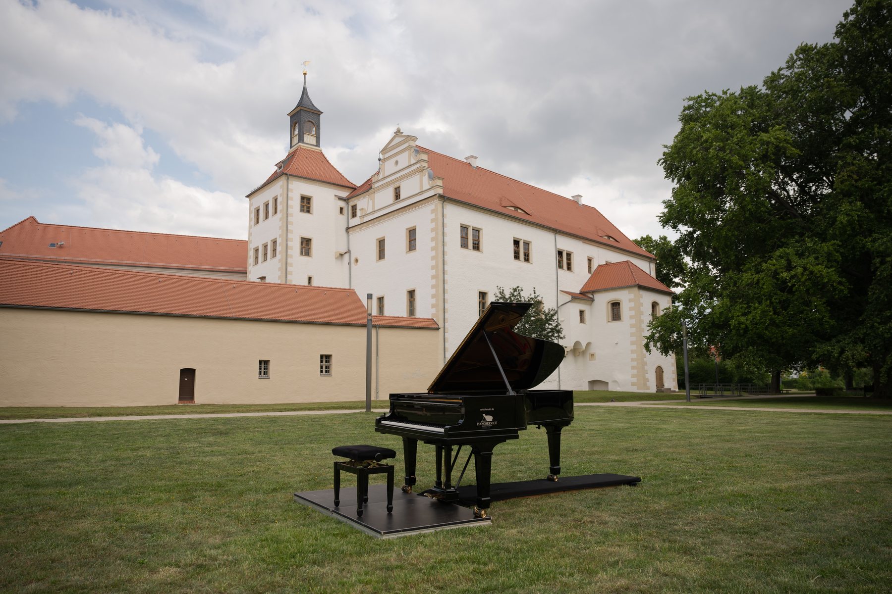 Ein offener Konzertflügel auf einer Wiese vor einem weißen Schloss mit roten Dächern