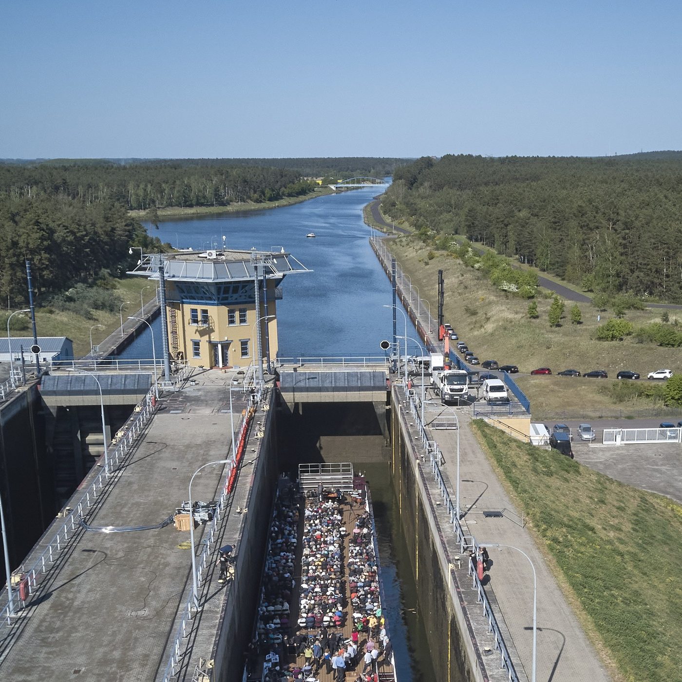 Luftbild: das voll besetzte Schiff liegt tief unten in der Schleusenkammer, dahinter der Kanal und Kiefernwald