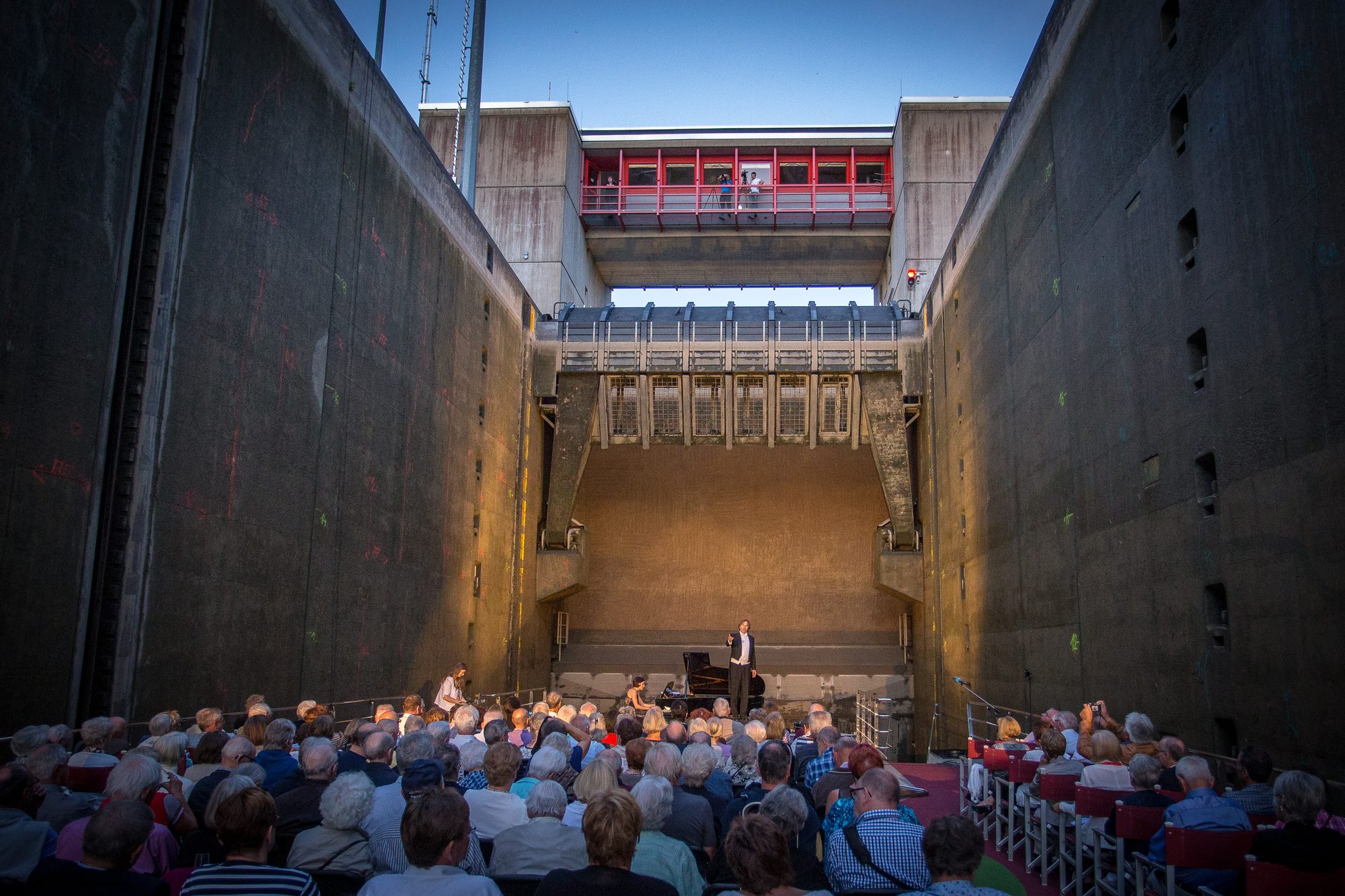 Voll besetztes Deck in der Schleusenkammer vor dem golden angestrahlten Schleusentor, vorne Sänger und Pianistin