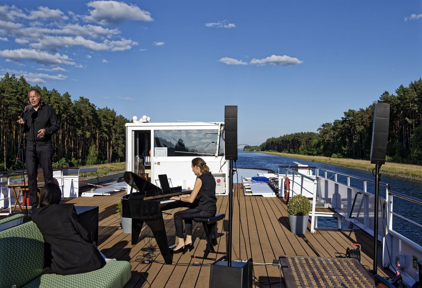 Sänger und Pianistin an Deck eines Flussschiffes, während Kanal und Kiefernwald vorbeiziehen