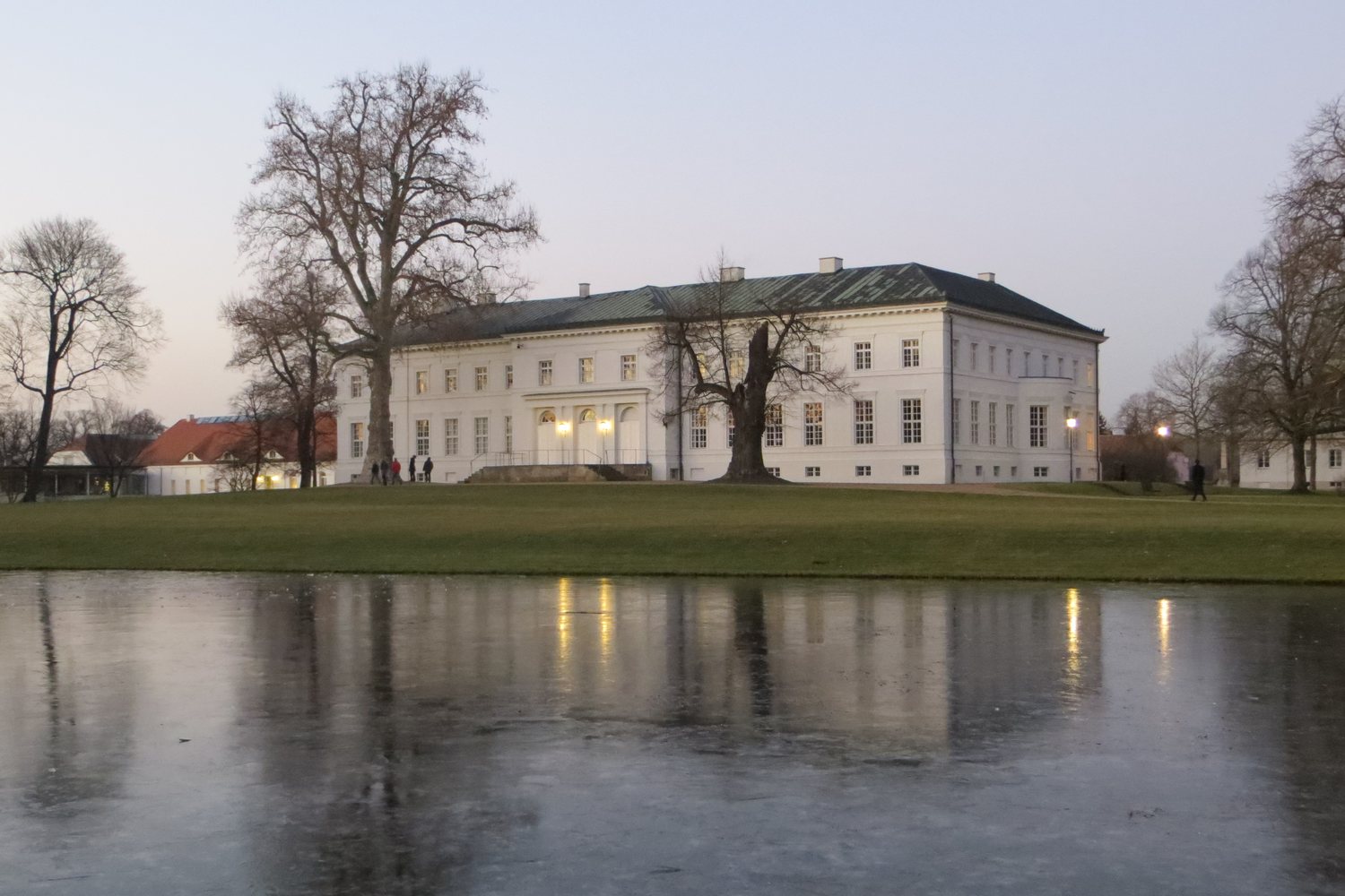Schloss Neuhardenberg in der Winterdämmerung, davor der zugefrorene Teich mit Spiegelung der erleuchteten Fenster
