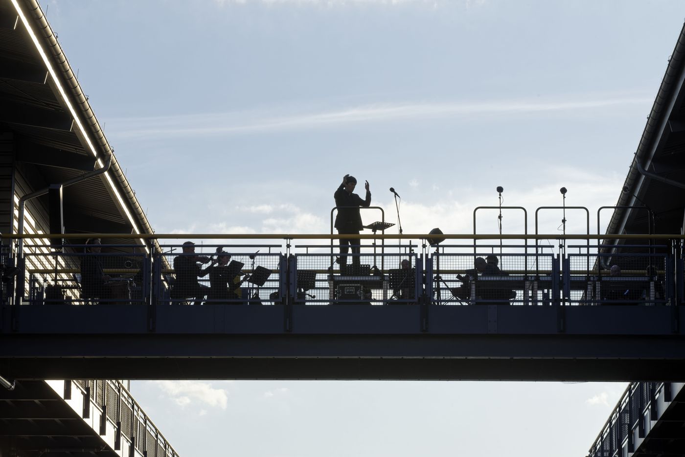 Ein Dirigent als Scherenschnitt auf der Brücke des Schiffshebewerks Niederfinow vor hellem Himmel, darunter das Ensemble